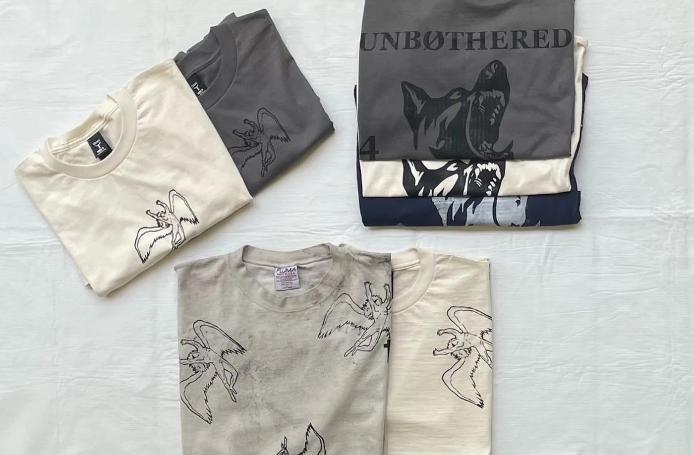 Original photograph of folded Wings and Unbothered tees in cream, gray and navy