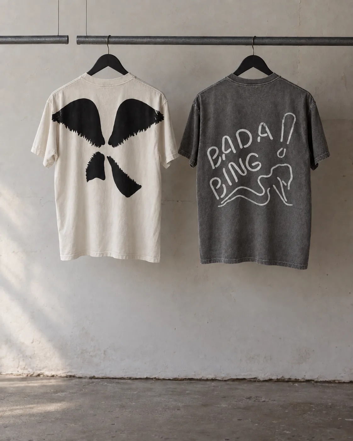 Studio visualization of cream Wings and gray Bada Bing back-print tees hanging on a steel rack